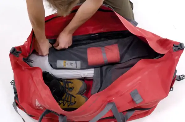 a red holdall that is fully open showing camping gear as its contents. A mands hands are visible closing a zip