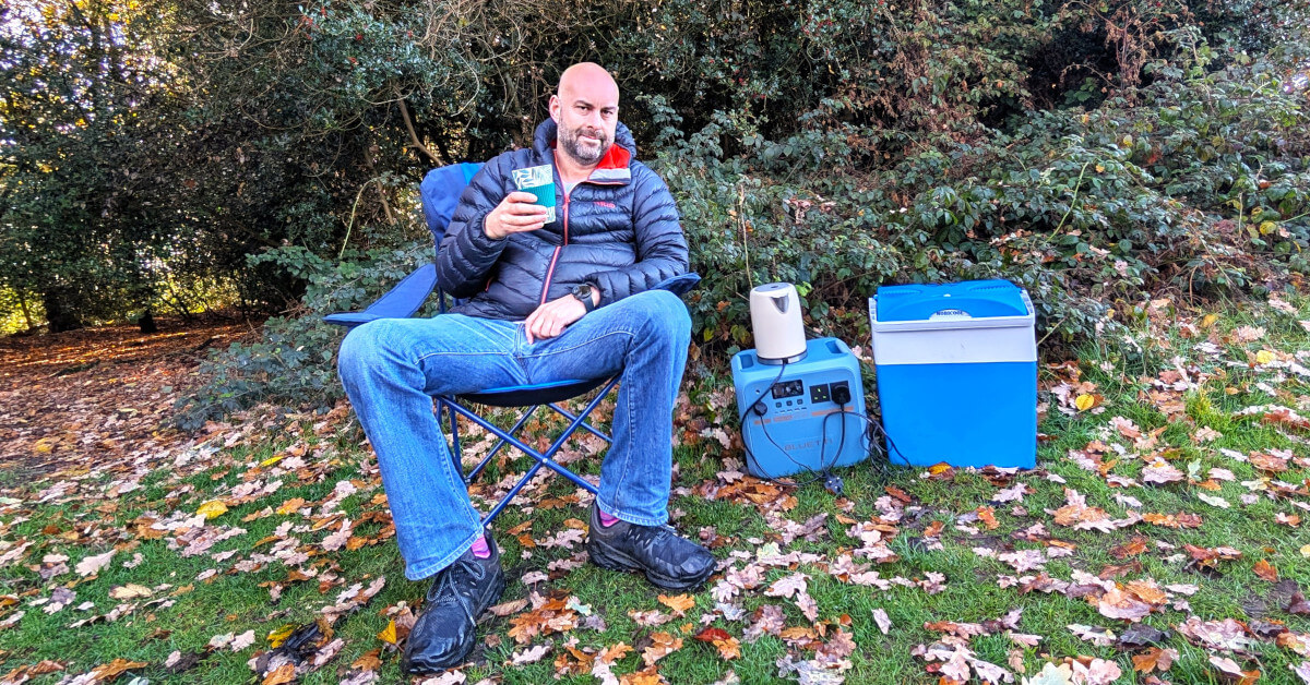 a man sitting in a camping chair with a Bluetti Pioneer Na power station next to him powering a kettle and coolbox